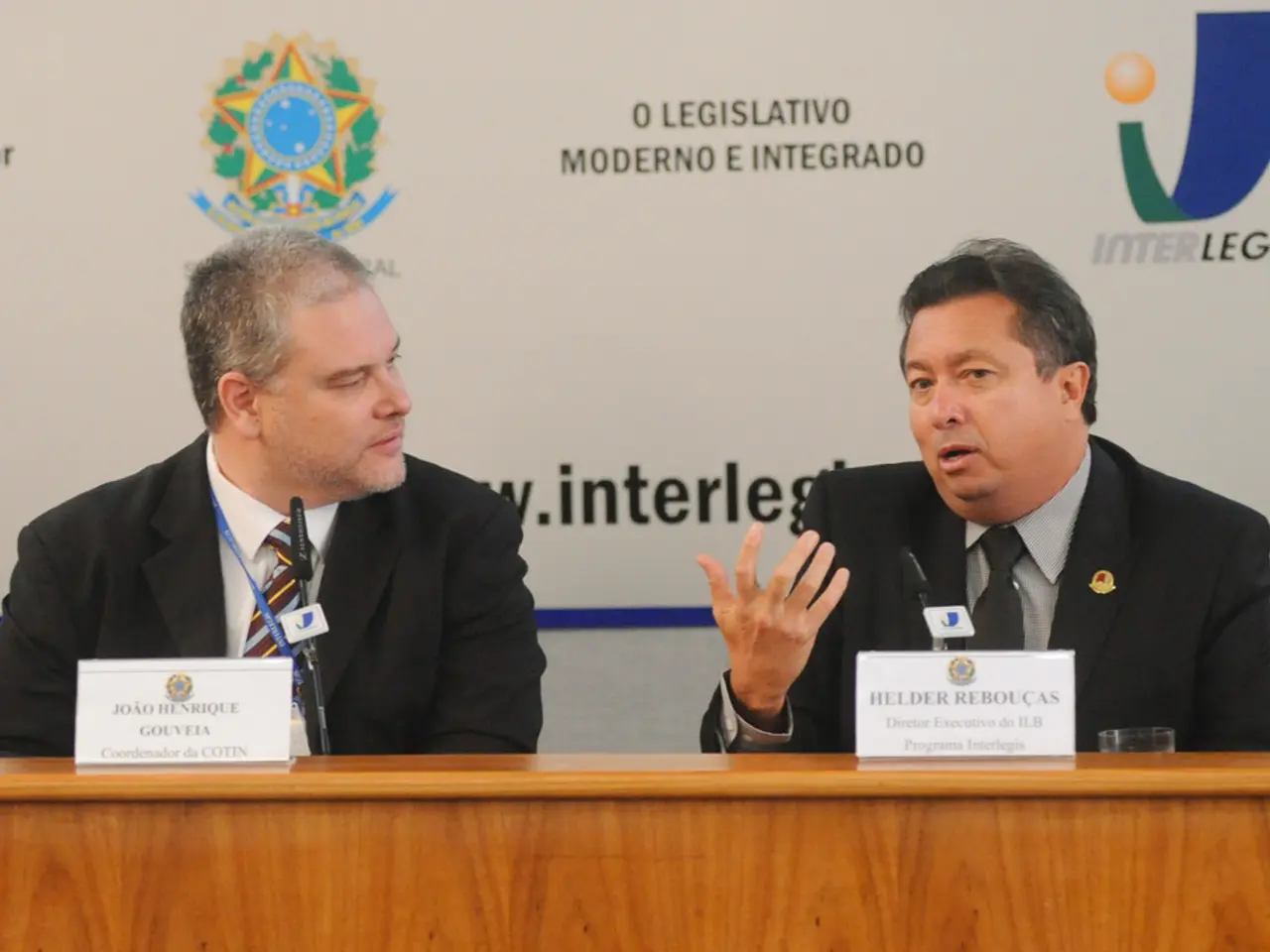 In this image I see 2 men who are wearing suits and I see a table in front of them on which there...