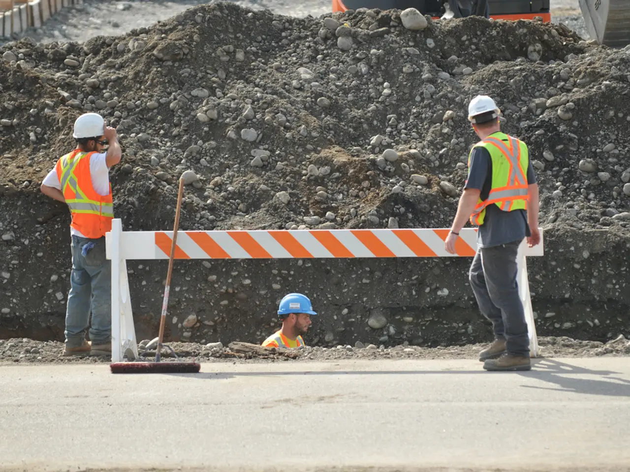 In this image there are people standing on a road and a person digging soil, in the background...