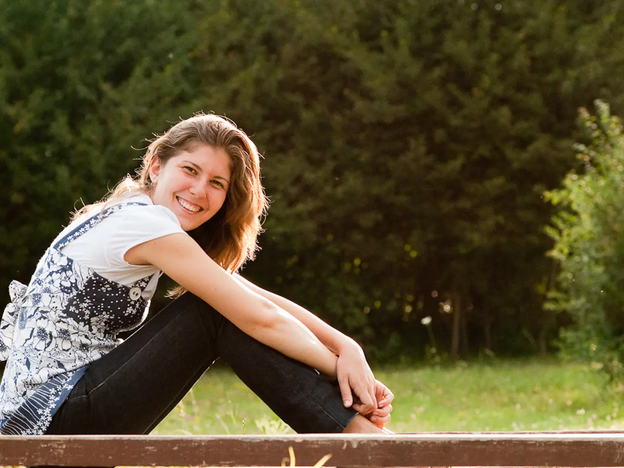 This is the woman sitting and smiling. She wore a top and trouser. This looks like a wood. Here is...