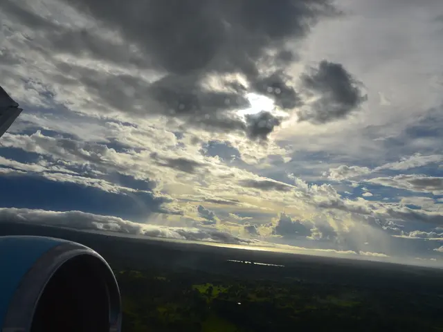 This image has taken from an aeroplane where we can see sky with clouds and land with greenery. At...