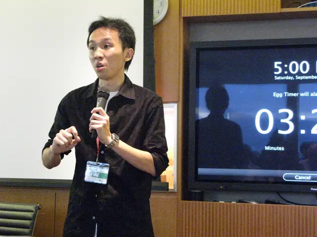 There is a man standing and holding a microphone and talking and wore tag,behind this man we can...