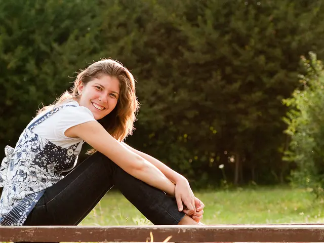 This is the woman sitting and smiling. She wore a top and trouser. This looks like a wood. Here is...