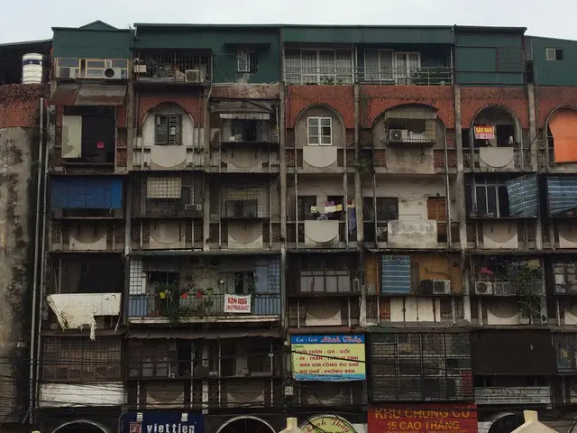 Picture of a building. Here we can see windows, plants, hoardings, banners, grill and sky.